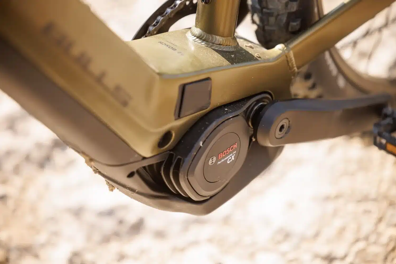 Nahaufnahme des schwarzen Bosch Performance Line CX Motors, der am Tretlager eines goldfarbenen BULLS E-Mountainbikes montiert ist. Kurbelarm und Teile des Rahmens sind sichtbar.