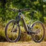 Bulls Copperhead EVO AM 2 E-Mountainbike in olivgrüner Lackierung, mit Federgabel und voller Federung, steht auf einem Feldweg im Grünen.