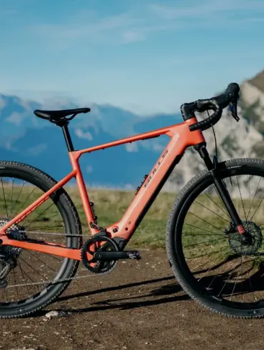 Dynamische Ansicht des BULLS Machete EVO SX 1 E-Gravelbikes, das auf einem Schotterweg mit Bergkulisse im Hintergrund steht. Das Fahrrad ist orange und zeigt seine sportlichen Komponenten.