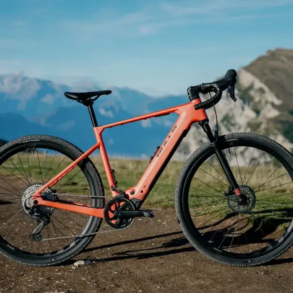 Dynamische Ansicht des BULLS Machete EVO SX 1 E-Gravelbikes, das auf einem Schotterweg mit Bergkulisse im Hintergrund steht. Das Fahrrad ist orange und zeigt seine sportlichen Komponenten.