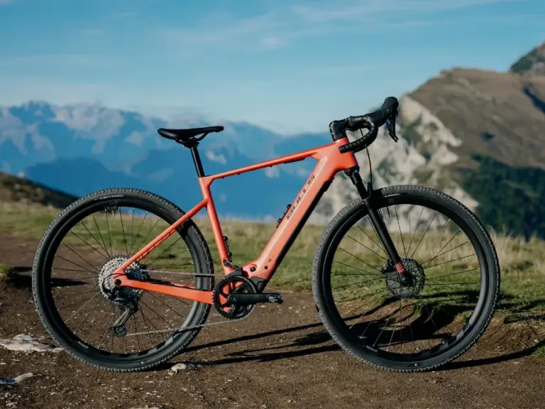 Dynamische Ansicht des BULLS Machete EVO SX 1 E-Gravelbikes, das auf einem Schotterweg mit Bergkulisse im Hintergrund steht. Das Fahrrad ist orange und zeigt seine sportlichen Komponenten.