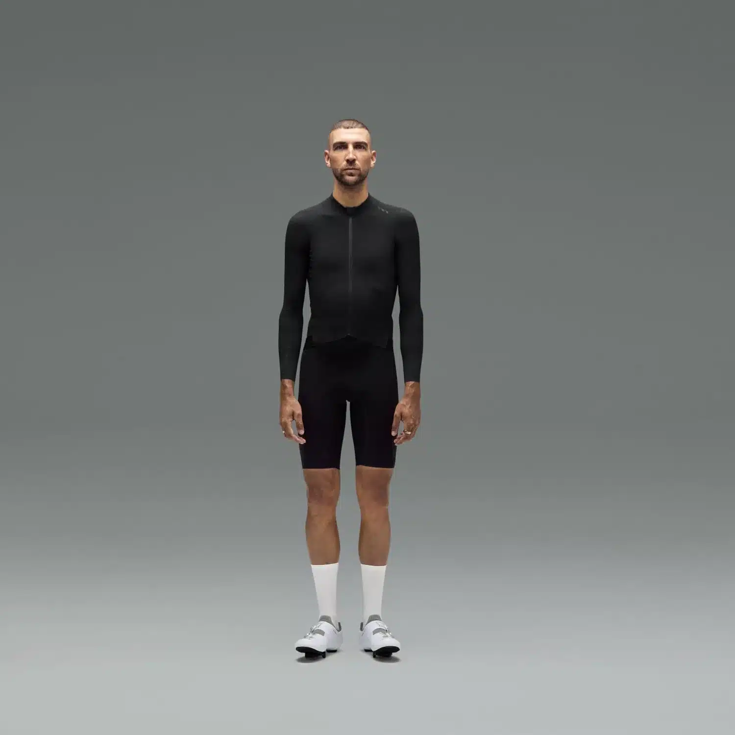 Mann mit sportlicher Figur steht frontal in einem schwarzen Langarm-Radtrikot, schwarzen Radhosen, weißen Socken und weißen Radschuhen vor grauem Hintergrund.