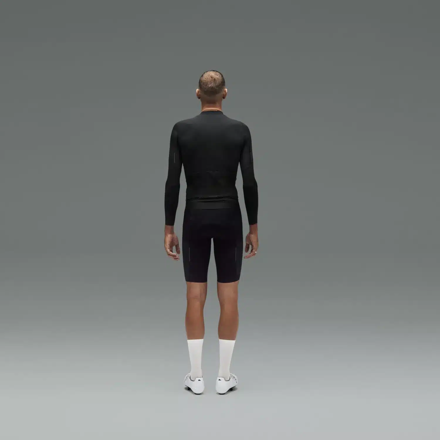 Rückansicht eines Mannes mit kurzgeschorenem Haar, der ein schwarzes Canyon CFR Langarm-Radtrikot und schwarze Radhosen mit weißen Socken und weißen Radschuhen trägt, vor einem neutralen grauen Hintergrund.