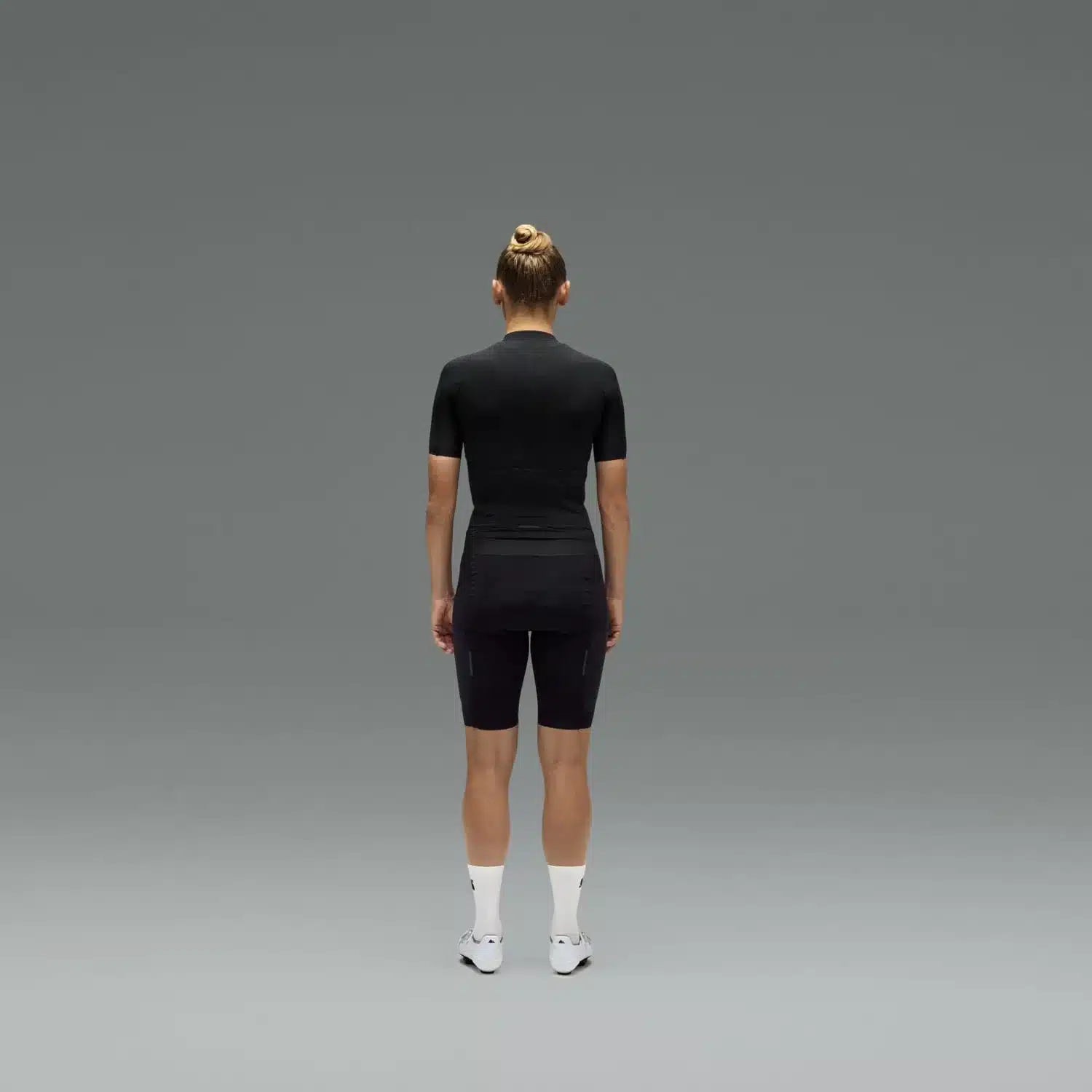 Rückansicht einer Frau, die ein schwarzes, kurzärmeliges Radsporttrikot und eine schwarze Radhose trägt, auf neutralem Hintergrund.
