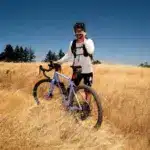 Mann mit violettem Canyon Grizl CF 9 w/ ECLIPS Gravelbike in einem trockenen Feld, blaue Himmel, Bäume am Horizont.