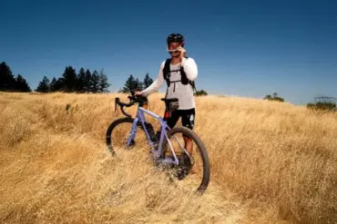 Mann mit violettem Canyon Grizl CF 9 w/ ECLIPS Gravelbike in einem trockenen Feld, blaue Himmel, Bäume am Horizont.