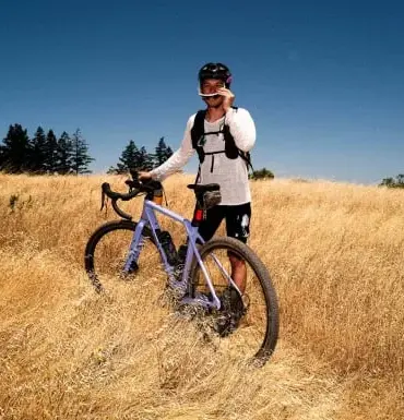 Mann mit violettem Canyon Grizl CF 9 w/ ECLIPS Gravelbike in einem trockenen Feld, blaue Himmel, Bäume am Horizont.