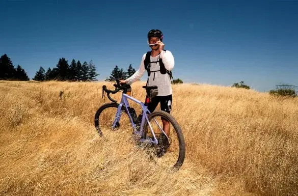 Mann mit violettem Canyon Grizl CF 9 w/ ECLIPS Gravelbike in einem trockenen Feld, blaue Himmel, Bäume am Horizont.