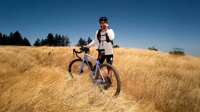 Mann mit violettem Canyon Grizl CF 9 w/ ECLIPS Gravelbike in einem trockenen Feld, blaue Himmel, Bäume am Horizont.
