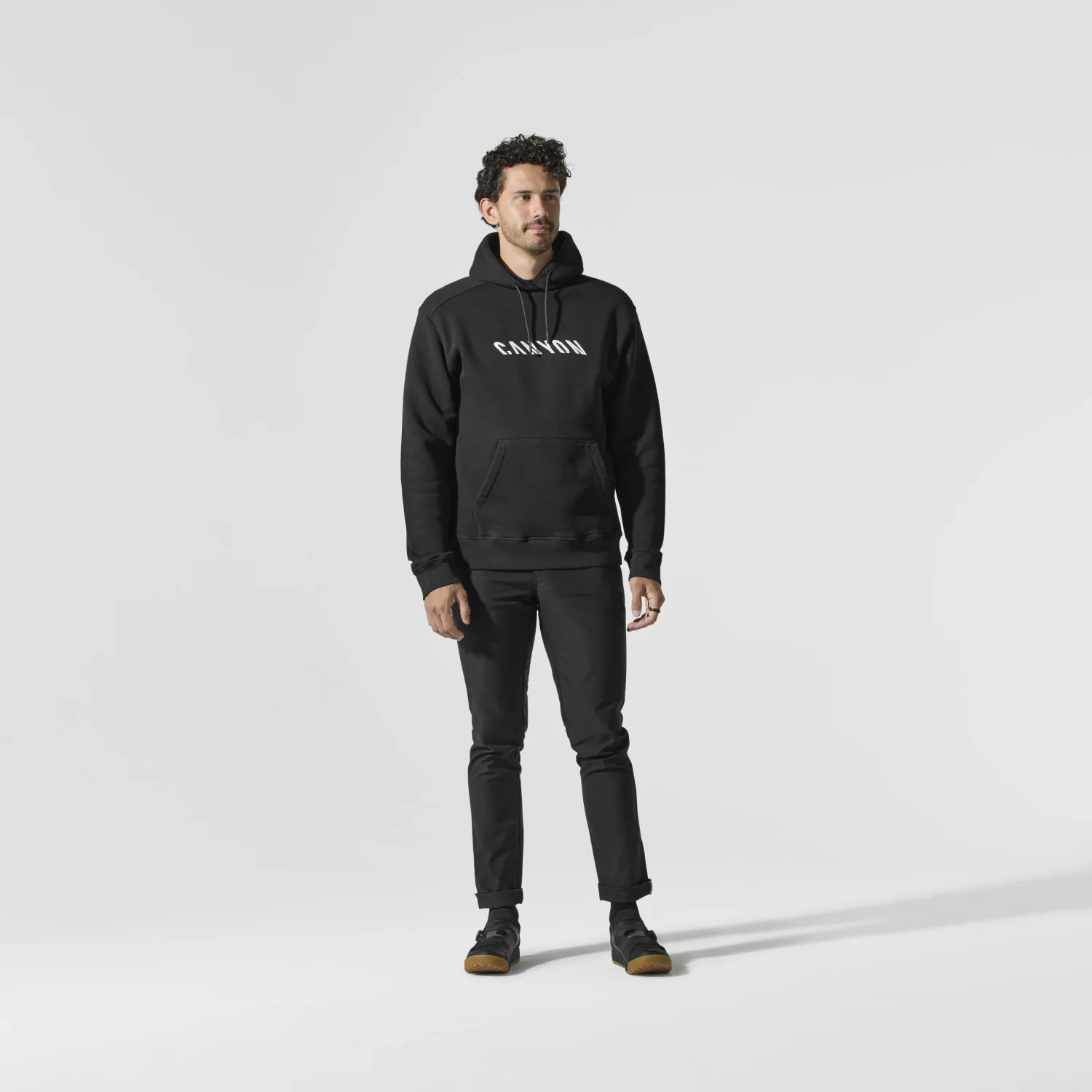 Ein Mann steht frontal im Bild und trägt einen schwarzen Canyon Hoodie mit weißem
