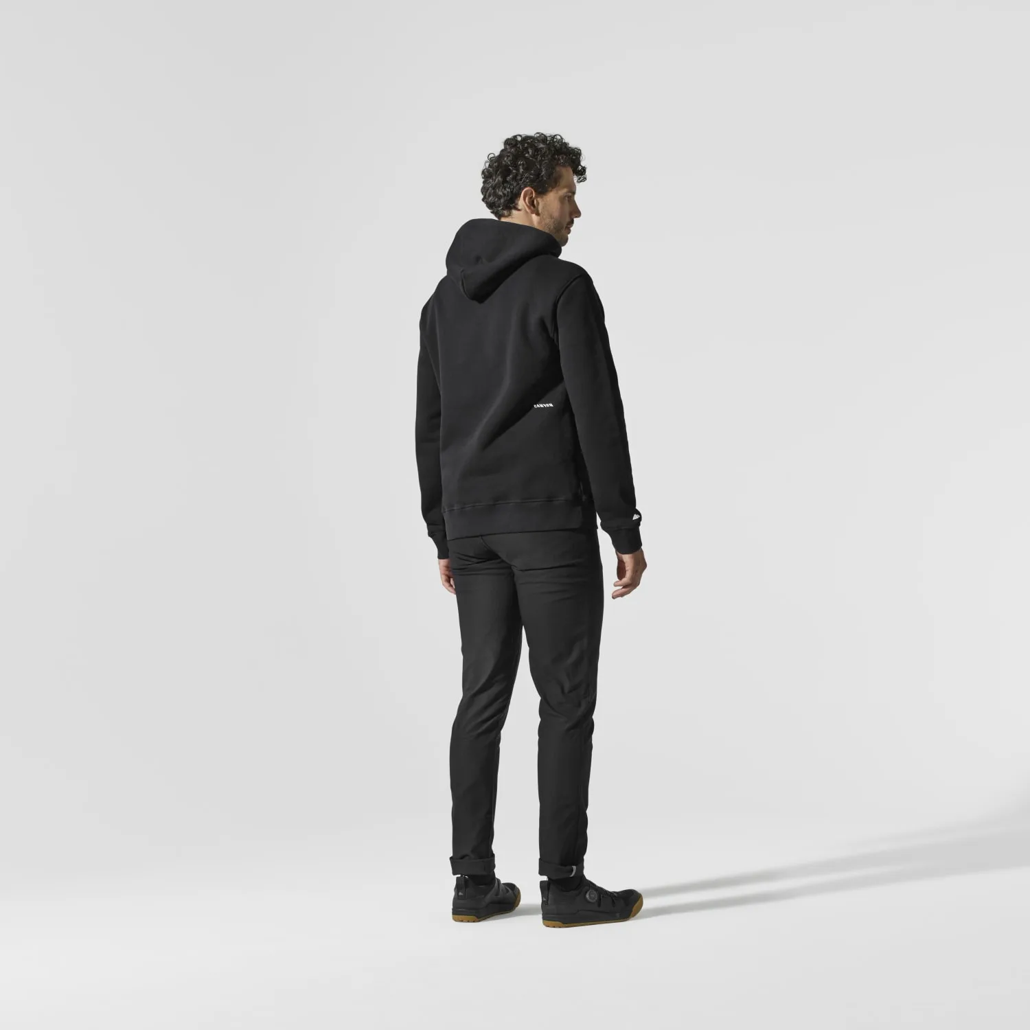 Rückenansicht eines Mannes, der einen schwarzen Kapuzenpullover von Canyon und passende schwarze Hose trägt, auf hellem Hintergrund.