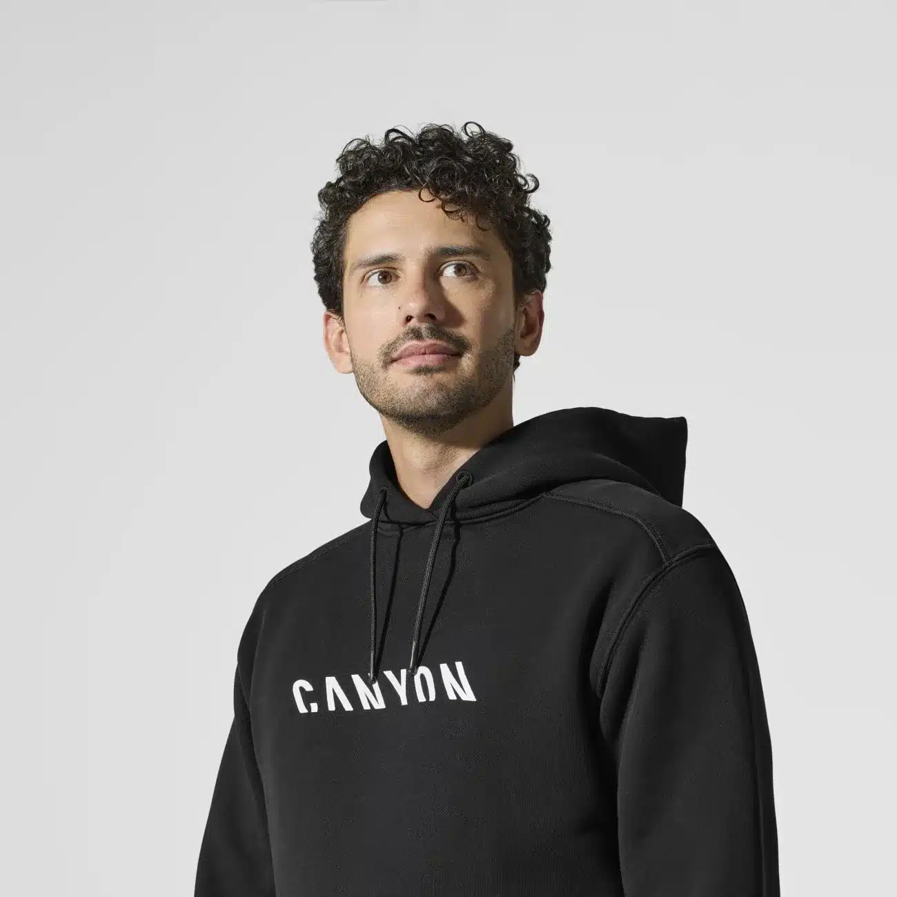 Ein Mann mit Locken und Bart trägt einen schwarzen Canyon Hoodie. Der Schriftzug Canyon ist in weißer Schrift auf der Vorderseite des Hoodies zu sehen.