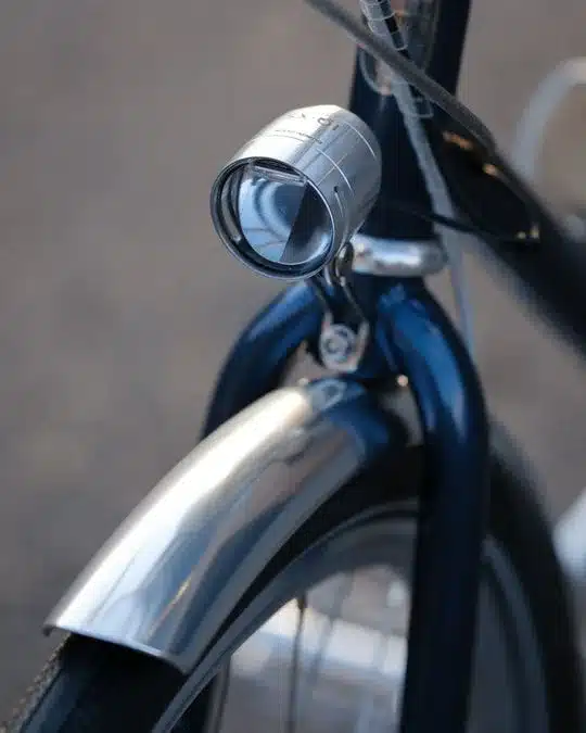 Nahaufnahme des vorderen Bereichs eines dunkelblauen Fahrrads mit einem glänzenden, runden Scheinwerfer und einem silbernen Schutzblech über dem Vorderrad.