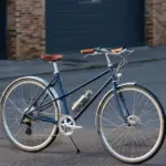 Dunkelblaues Capri Azur 4 E-Bike mit braunem Sattel und Griffen, silbernen Felgen und Schutzblechen, befestigter Trinkflasche, steht auf asphaltiertem Untergrund vor einer grauen Garagentür.