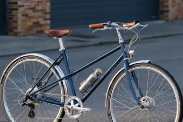 Dunkelblaues Capri Azur 4 E-Bike mit braunem Sattel und Griffen, silbernen Felgen und Schutzblechen, befestigter Trinkflasche, steht auf asphaltiertem Untergrund vor einer grauen Garagentür.