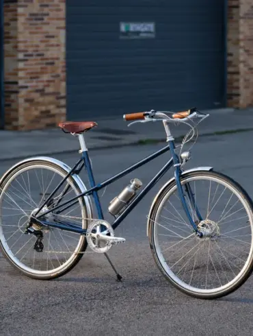 Dunkelblaues Capri Azur 4 E-Bike mit braunem Sattel und Griffen, silbernen Felgen und Schutzblechen, befestigter Trinkflasche, steht auf asphaltiertem Untergrund vor einer grauen Garagentür.