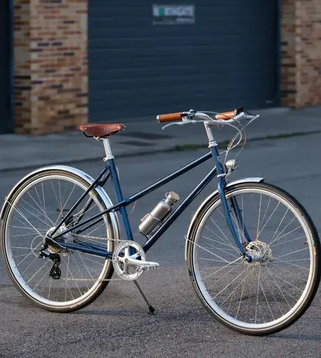 Dunkelblaues Capri Azur 4 E-Bike mit braunem Sattel und Griffen, silbernen Felgen und Schutzblechen, befestigter Trinkflasche, steht auf asphaltiertem Untergrund vor einer grauen Garagentür.