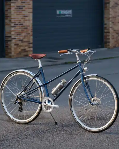 Dunkelblaues Capri Azur 4 E-Bike mit braunem Sattel und Griffen, silbernen Felgen und Schutzblechen, befestigter Trinkflasche, steht auf asphaltiertem Untergrund vor einer grauen Garagentür.