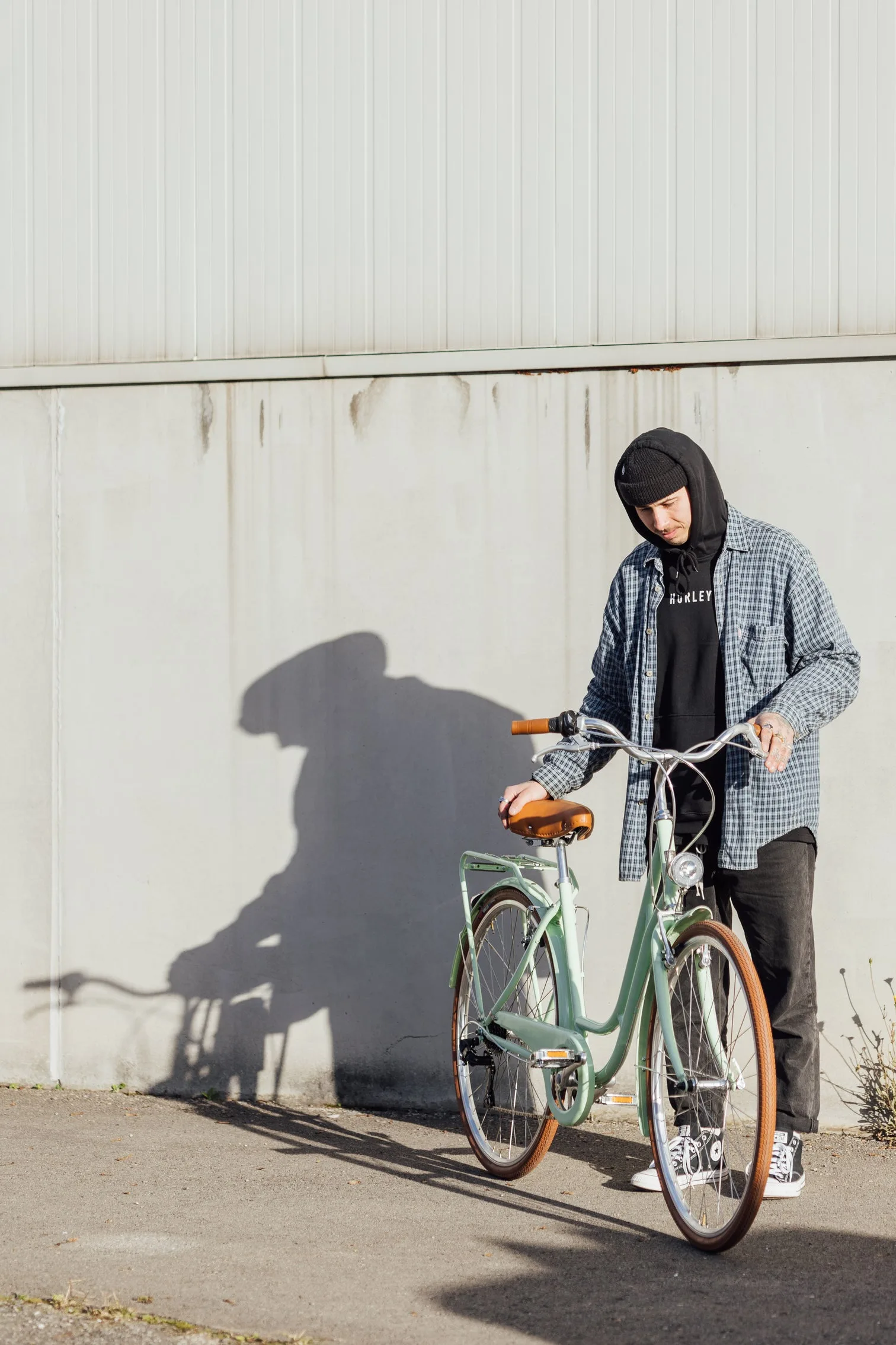 Ein Mann in legerer Kleidung steht neben einem mintgrünen Capri Berlin Fahrrad, das er zu betrachten scheint. Der Hintergrund ist eine helle, vertikal geriffelte Wand mit einer Schattenprojektion des Mannes und des Fahrrads.