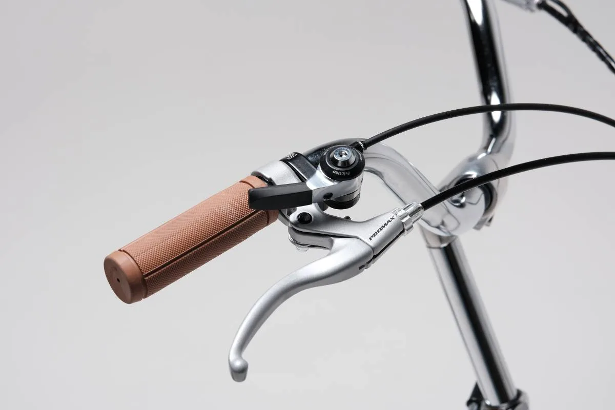 Detailansicht des Lenkers eines silbernen Retro-Faltrades mit einem braunen Griff und einem silbernen Promax Bremshebel, Bowdenzüge und Schalthebel sind sichtbar.