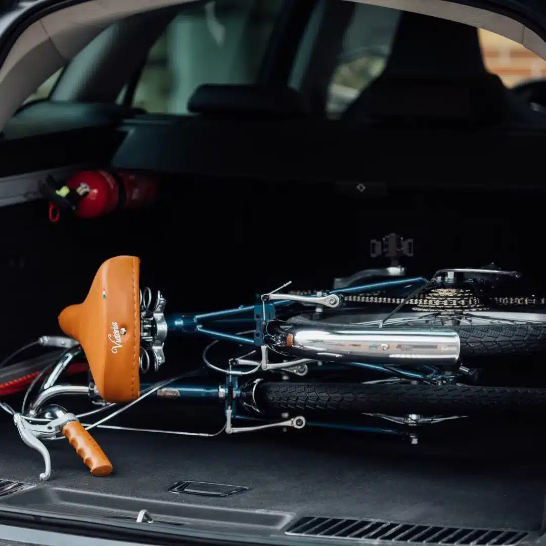 Dunkelblaues Capri Vita Falt-E-Bike in zusammengeklapptem Zustand, liegend im dunklen Kofferraum eines Autos. Ein brauner Ledersattel mit der Aufschrift