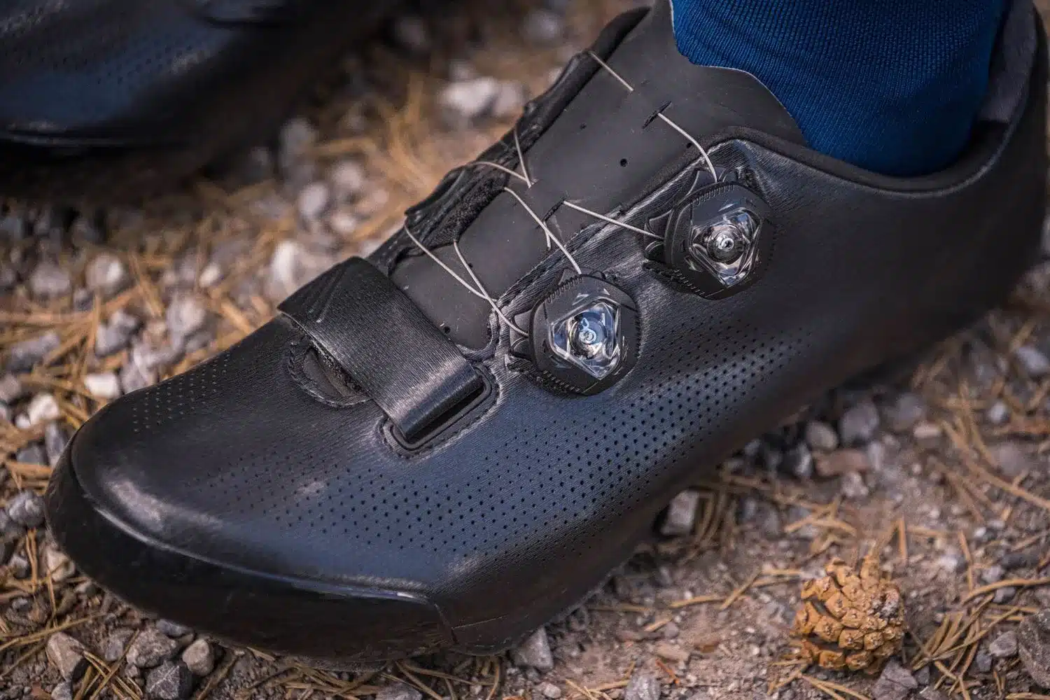 Nahaufnahme eines schwarzen Charge Pro MTB-Schuhs mit zwei BOA-Drehverschlüssen und einem Klettverschluss, der auf steinigem Boden liegt.