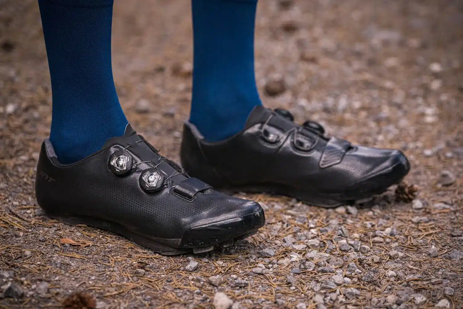 Schwarze MTB-Fahrradschuhe mit BOA-Verschlüssen und blauen Socken, aus der Nähe auf einem Feldweg fotografiert.