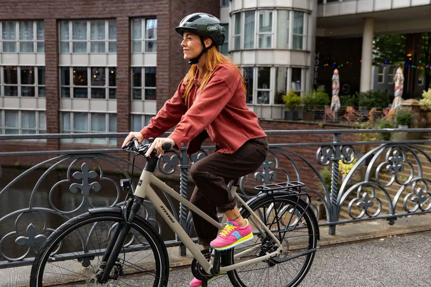 Frau mit Helm fährt ein beiges Trekkingrad vom Hersteller Stevens mit einem Gepäckträger über eine Brücke.