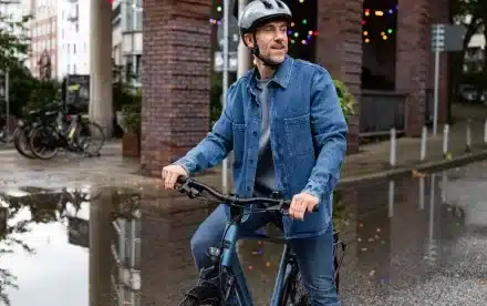 Mann mit Helm und Jeansjacke sitzt auf einem blauen Tourenrad, das auf nasser Straße steht.