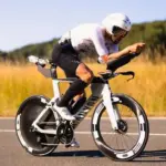 Triathlet mit Helm und Brille fährt ein weißes Canyon Speedmax CFR 1by AXS Rennrad auf asphaltierter Straße, umgeben von Feldern und Bäumen.