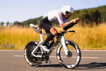 Triathlet mit Helm und Brille fährt ein weißes Canyon Speedmax CFR 1by AXS Rennrad auf asphaltierter Straße, umgeben von Feldern und Bäumen.