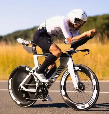 Triathlet mit Helm und Brille fährt ein weißes Canyon Speedmax CFR 1by AXS Rennrad auf asphaltierter Straße, umgeben von Feldern und Bäumen.