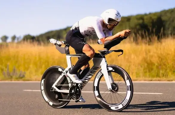 Triathlet mit Helm und Brille fährt ein weißes Canyon Speedmax CFR 1by AXS Rennrad auf asphaltierter Straße, umgeben von Feldern und Bäumen.