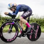 Radfahrerin in voller Rennausrüstung mit USA-Trikot auf einem schwarzen Canyon Speedmax CFR TT Zeitfahrrad, das auf einer asphaltierten Straße fährt.