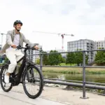 Ein Mann in Freizeitkleidung fährt ein schwarzes E-Triton 9.8.1 Plus HT Fahrrad auf einem Uferweg mit modernen Gebäuden im Hintergrund.