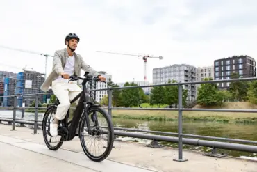 Ein Mann in Freizeitkleidung fährt ein schwarzes E-Triton 9.8.1 Plus HT Fahrrad auf einem Uferweg mit modernen Gebäuden im Hintergrund.