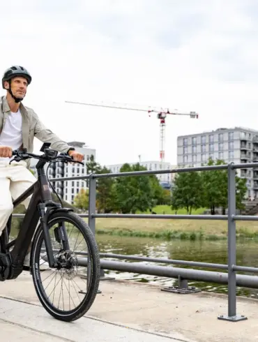 Ein Mann in Freizeitkleidung fährt ein schwarzes E-Triton 9.8.1 Plus HT Fahrrad auf einem Uferweg mit modernen Gebäuden im Hintergrund.