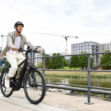 Ein Mann in Freizeitkleidung fährt ein schwarzes E-Triton 9.8.1 Plus HT Fahrrad auf einem Uferweg mit modernen Gebäuden im Hintergrund.