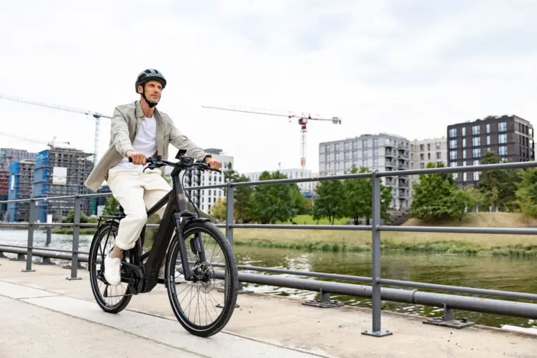Ein Mann in Freizeitkleidung fährt ein schwarzes E-Triton 9.8.1 Plus HT Fahrrad auf einem Uferweg mit modernen Gebäuden im Hintergrund.