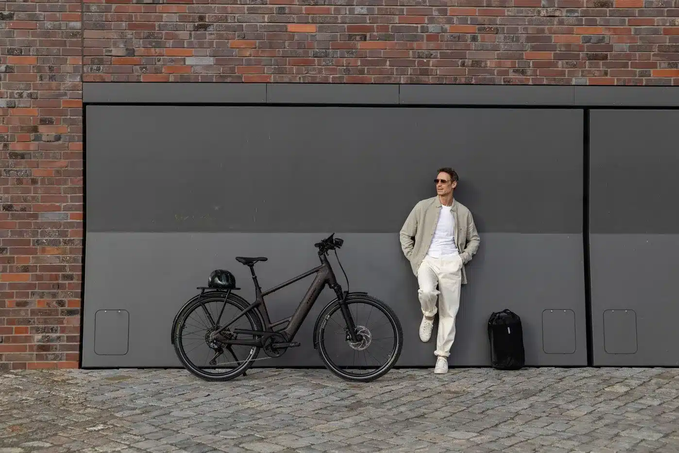 Ein Mann in Freizeitkleidung steht neben einem dunkelbraunen E-Triton 9.8.1 Plus HT Fahrrad, das mit Gepäckträger, Schutzblechen und einem Helm ausgestattet ist. Er lehnt an einer grauen, modernen Wand mit einer Ziegelwand im Hintergrund.