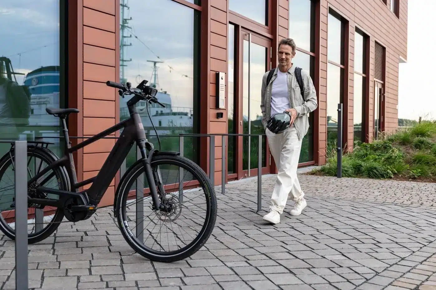Schwarzes E-Triton 9.8.1 Plus HT Touren-Pedelec mit Gepäckträger und Bosch-Motor, ein Mann mit Helm geht im Hintergrund.