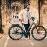 Ein Mann mit Helm steht neben einem grauen Giant AnyTour E+ 1 E-Bike auf einem asphaltierten Radweg. Er trägt ein helles Hemd und eine dunkle Hose. Im Hintergrund sind Bäume und eine Mauer mit Ranken zu sehen.