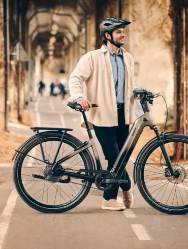 Ein Mann mit Helm steht neben einem grauen Giant AnyTour E+ 1 E-Bike auf einem asphaltierten Radweg. Er trägt ein helles Hemd und eine dunkle Hose. Im Hintergrund sind Bäume und eine Mauer mit Ranken zu sehen.
