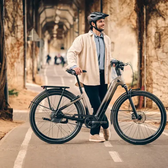 Ein Mann mit Helm steht neben einem grauen Giant AnyTour E+ 1 E-Bike auf einem asphaltierten Radweg. Er trägt ein helles Hemd und eine dunkle Hose. Im Hintergrund sind Bäume und eine Mauer mit Ranken zu sehen.