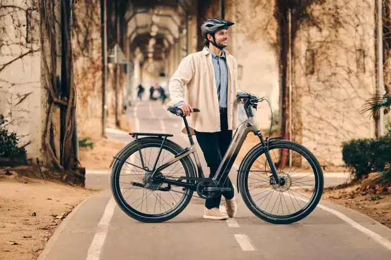 Ein Mann mit Helm steht neben einem grauen Giant AnyTour E+ 1 E-Bike auf einem asphaltierten Radweg. Er trägt ein helles Hemd und eine dunkle Hose. Im Hintergrund sind Bäume und eine Mauer mit Ranken zu sehen.