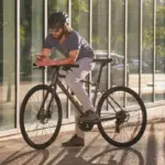 Mann mit Helm und Sonnenbrille auf einem dunklen Giant Escape 1 Fitnessbike stehend, mit einem Smartphone in der Hand.