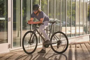 Mann mit Helm und Sonnenbrille auf einem dunklen Giant Escape 1 Fitnessbike stehend, mit einem Smartphone in der Hand.