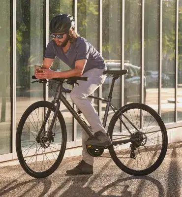 Mann mit Helm und Sonnenbrille auf einem dunklen Giant Escape 1 Fitnessbike stehend, mit einem Smartphone in der Hand.