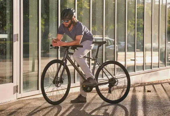 Mann mit Helm und Sonnenbrille auf einem dunklen Giant Escape 1 Fitnessbike stehend, mit einem Smartphone in der Hand.