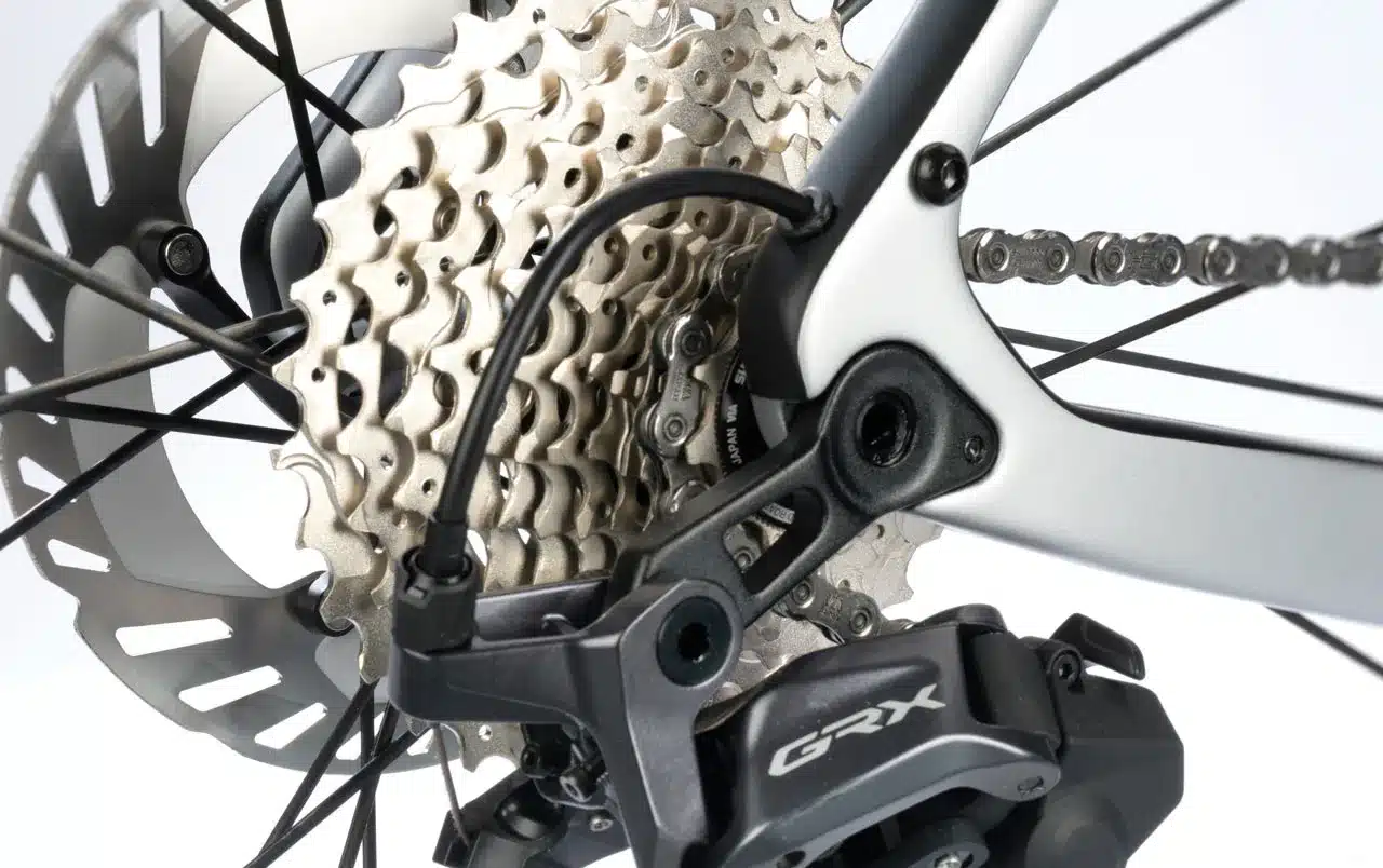 Detailaufnahme eines modernen Fahrrad-Antriebsstrangs mit Fokus auf das Schaltwerk, die Kette und die Kassette, neben einer polierten Bremsscheibe.